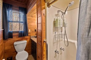 A wooden cabin bathroom with a toilet, a shower with a glass enclosure, and a window with curtains.