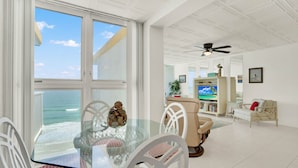 A dining area with a glass table and white chairs, a television, and a view of the ocean.