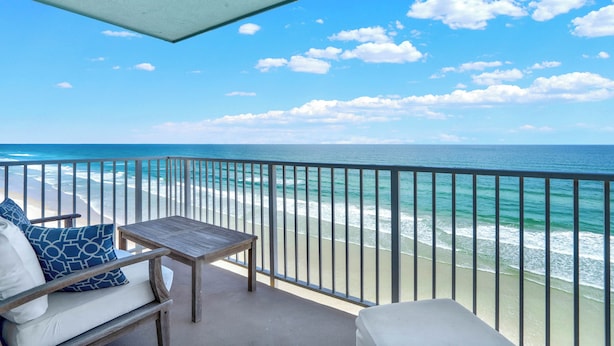 A balcony with a view of the ocean, a wooden table, and a chair with cushions.