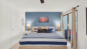 A bedroom with a bed, two bedside tables with lamps, a ceiling fan, a wall-mounted painting, and a sliding glass door leading to a balcony.