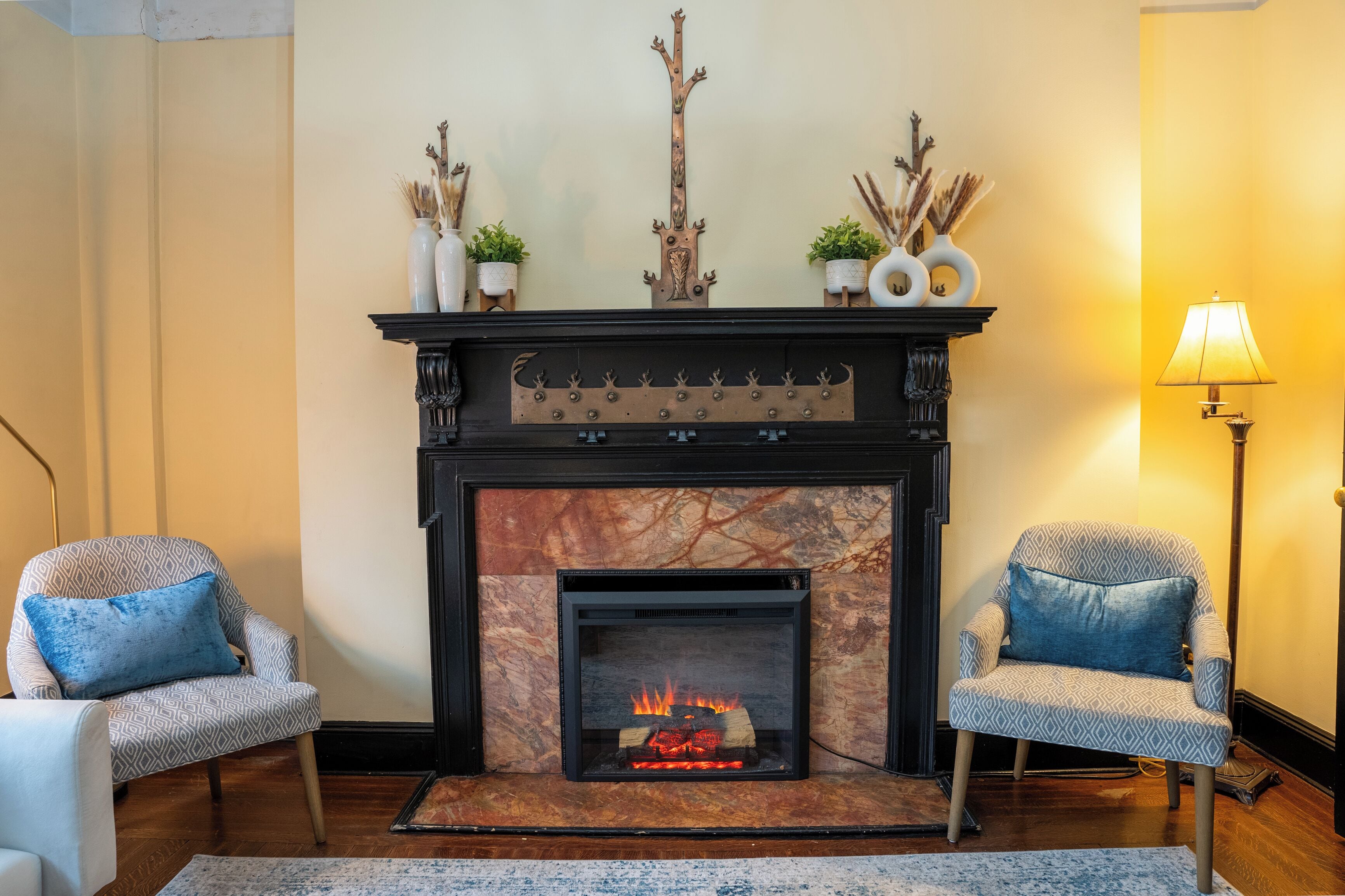 A living room with a fireplace, two patterned armchairs, a floor lamp, and decorative items on the mantel.