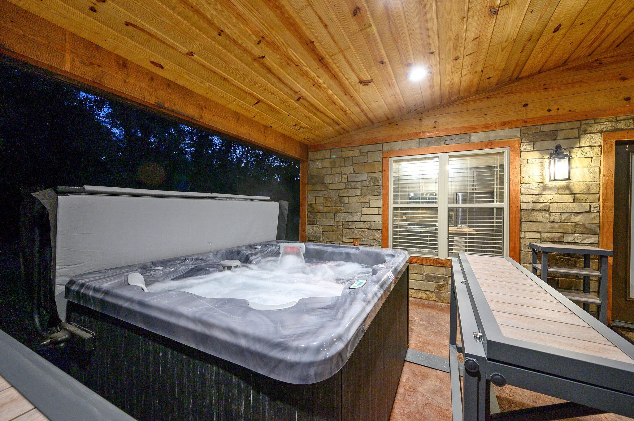 A hot tub in a wooden cabin with a stone wall and a window with blinds.