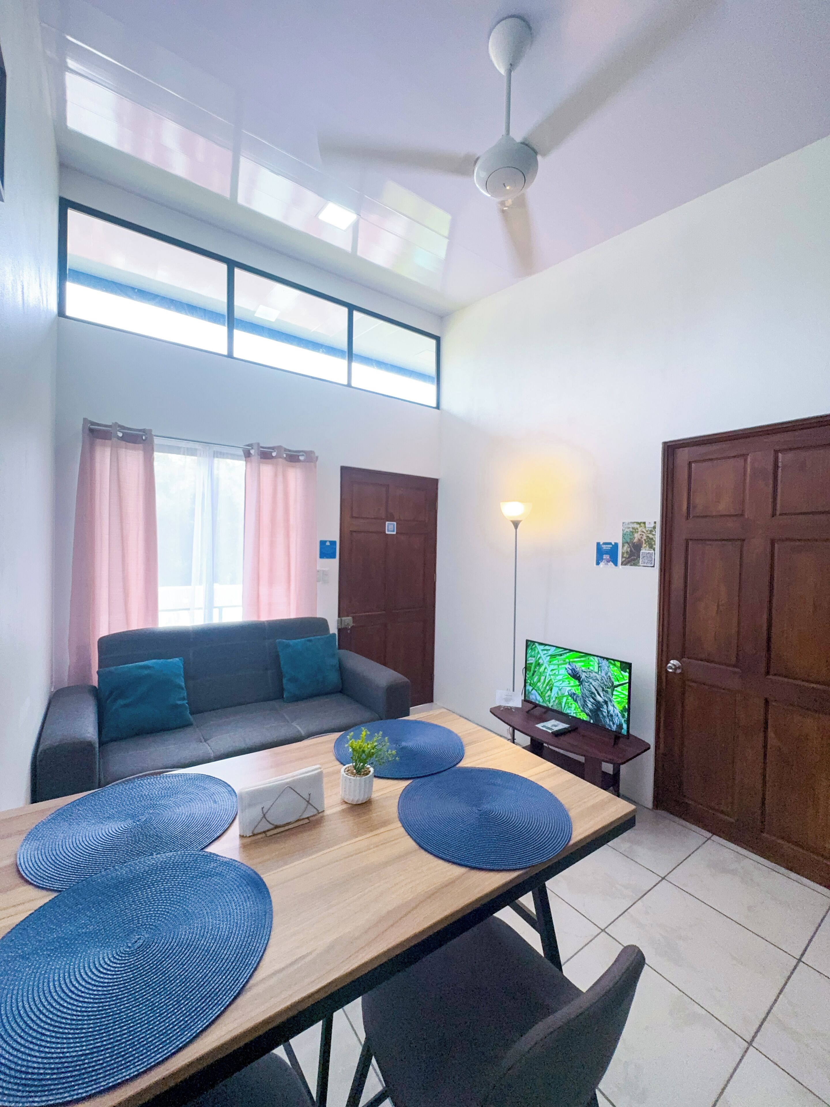 A modern living room with a sofa, a dining table with placemats, a television, and a ceiling fan.