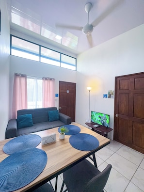 A modern living room with a sofa, a dining table with placemats, a television, and a ceiling fan.
