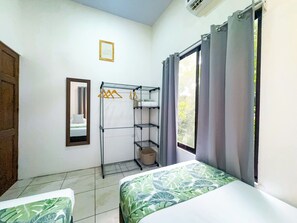 Une chambre d’hôtel comprenant un lit, une armoire, un miroir, une fenêtre avec des rideaux et une petite étagère avec des cintres.