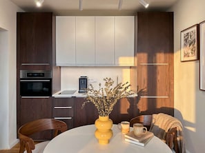 Eine moderne Küche mit dunklen Holzschränken, einer weißen Arbeitsplatte und einem eingebauten Backofen. Auf der Arbeitsplatte steht eine Vase mit Trockenblumen und eine Kaffeemaschine.