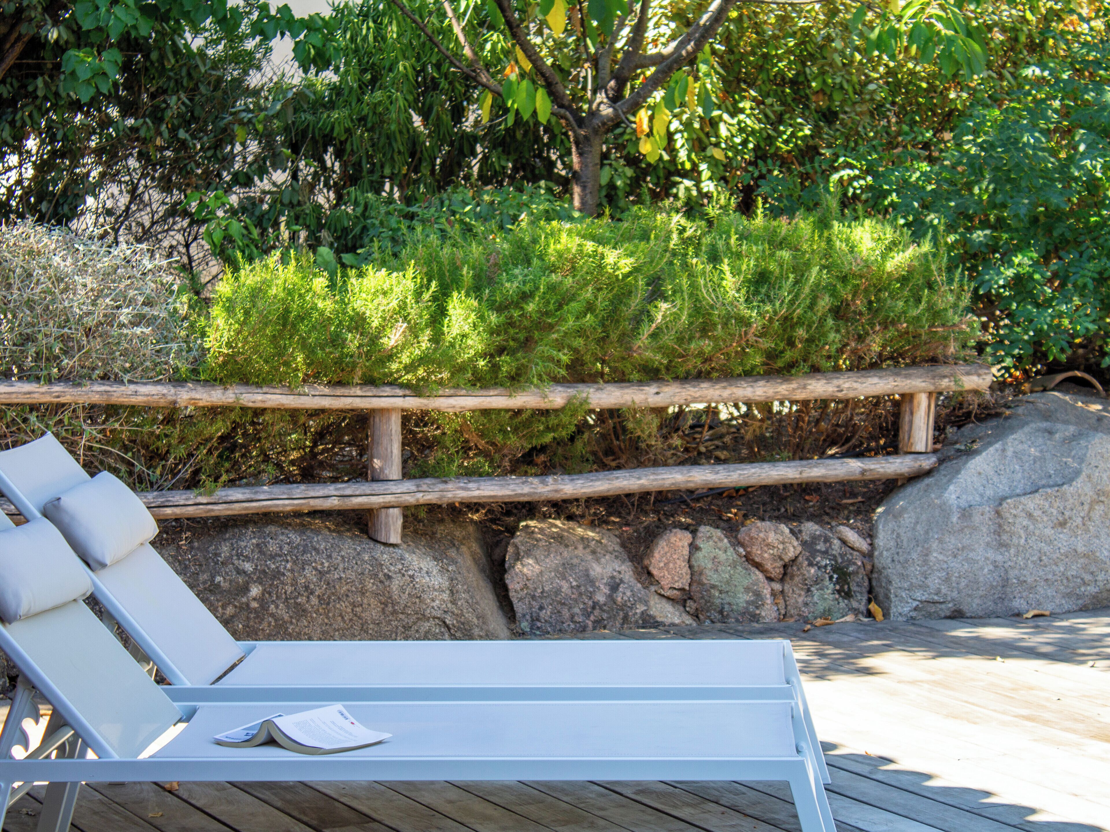 A wooden deck with a white lounge chair and a cushion, surrounded by greenery and rocks.