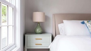 Quarto com cama, mesa de cabeceira com abajur, janela e piso de madeira.