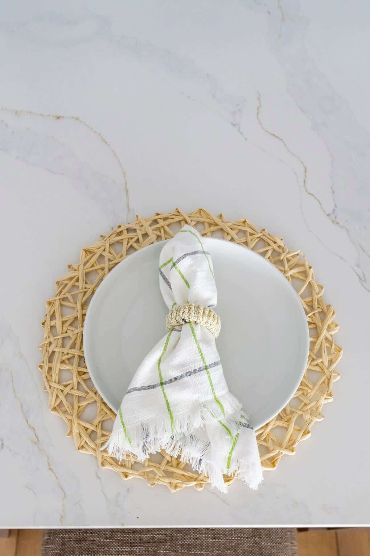 A white plate with a woven placemat, a white cloth with green stripes, and a beaded accessory on a marble surface.