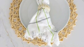 Um prato branco com um jogo de mantilha tecido, um pano branco com listras verdes e um adorno de contas sobre uma superfície de mármore.