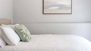 A neatly made bed with white bedding and decorative pillows, a framed artwork on the wall, and a plain white wall.