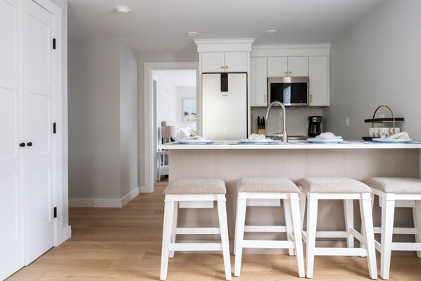 A modern kitchen with white cabinetry, a stainless steel refrigerator, a microwave, and a coffee machine. There are three white bar stools with cushioned seats.