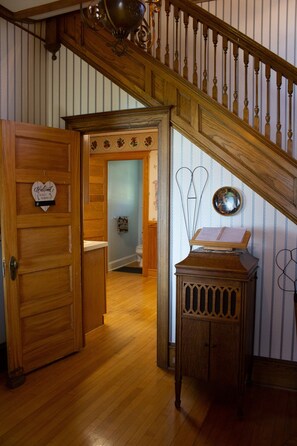 Una escalera de madera con barandilla decorativa que lleva a un rellano del segundo piso. Se ve una puerta de madera con un cartel de bienvenida con forma de corazón a la izquierda.