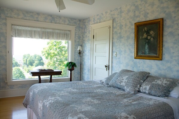 A bedroom with a large bed, a window with a view of trees, a door, and a wall-mounted painting.