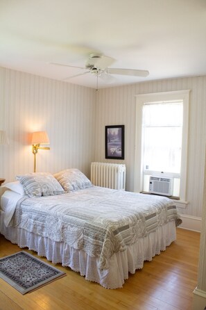 Une chambre avec un grand lit, un ventilateur de plafond, un radiateur, une fenêtre avec des stores et une lampe fixée au mur.