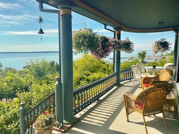 A covered outdoor area with wicker furniture, hanging flower baskets, and a view of the water. - Harbor Hill House Bayfield, WI (Bayfield)