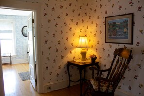 A room with floral wallpaper, a wooden chair, a table with a lamp, and a framed picture on the wall.