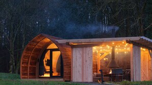 A cozy, semi-open wooden structure with a curved roof, a visible interior with a fireplace, and string lights. - Luxury Rural Cabin with Wood Fired Hot Tub (Canonbie)