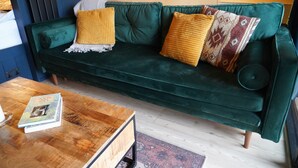 A green sofa with two yellow and one patterned cushion, a wooden coffee table with books, and a patterned rug. - Luxury Rural Cabin with Wood Fired Hot Tub (Canonbie)