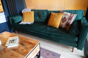 A green sofa with two yellow and one patterned cushion, a wooden coffee table with books, and a patterned rug.