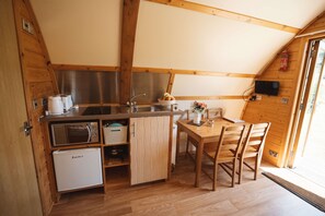 A compact kitchen with wooden cabinetry, a refrigerator, a microwave, and a sink. There is a dining table with chairs and a small window with a view.