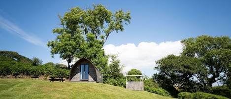 Eine kleine Hütte mit geneigtem Dach, umgeben von üppiger Vegetation und Bäumen, auf einem grasbewachsen Hügel.
