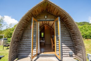 A wooden cabin with a pitched roof, a visible interior with a bed and a small kitchenette, and a door leading into the cabin. - Cosy Rural Cabin With Hot Tub (Pwllheli)