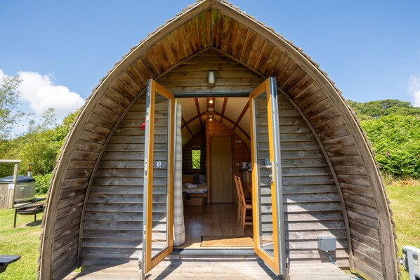 A wooden cabin with a pitched roof, a visible interior with a bed and a small kitchenette, and a door leading into the cabin.