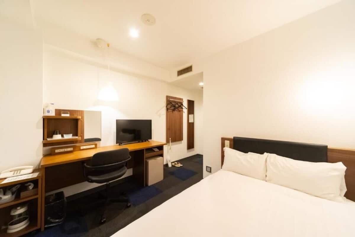 A hotel room with a bed, a desk with a computer, a chair, a TV, and a small kitchenette.