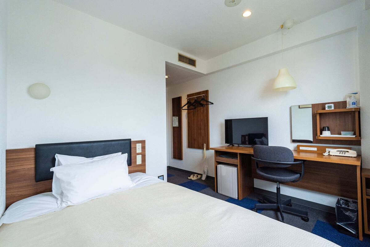 A hotel room with a bed, a desk with a chair, a TV, and a small refrigerator.