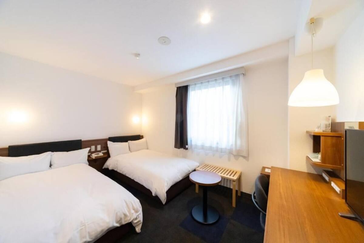 A hotel room with two beds, a desk, a chair, a small table, a TV, and a window with curtains.