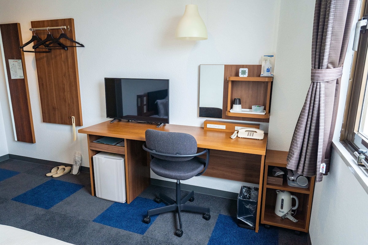 A hotel room with a desk, chair, TV, and a small kitchenette.