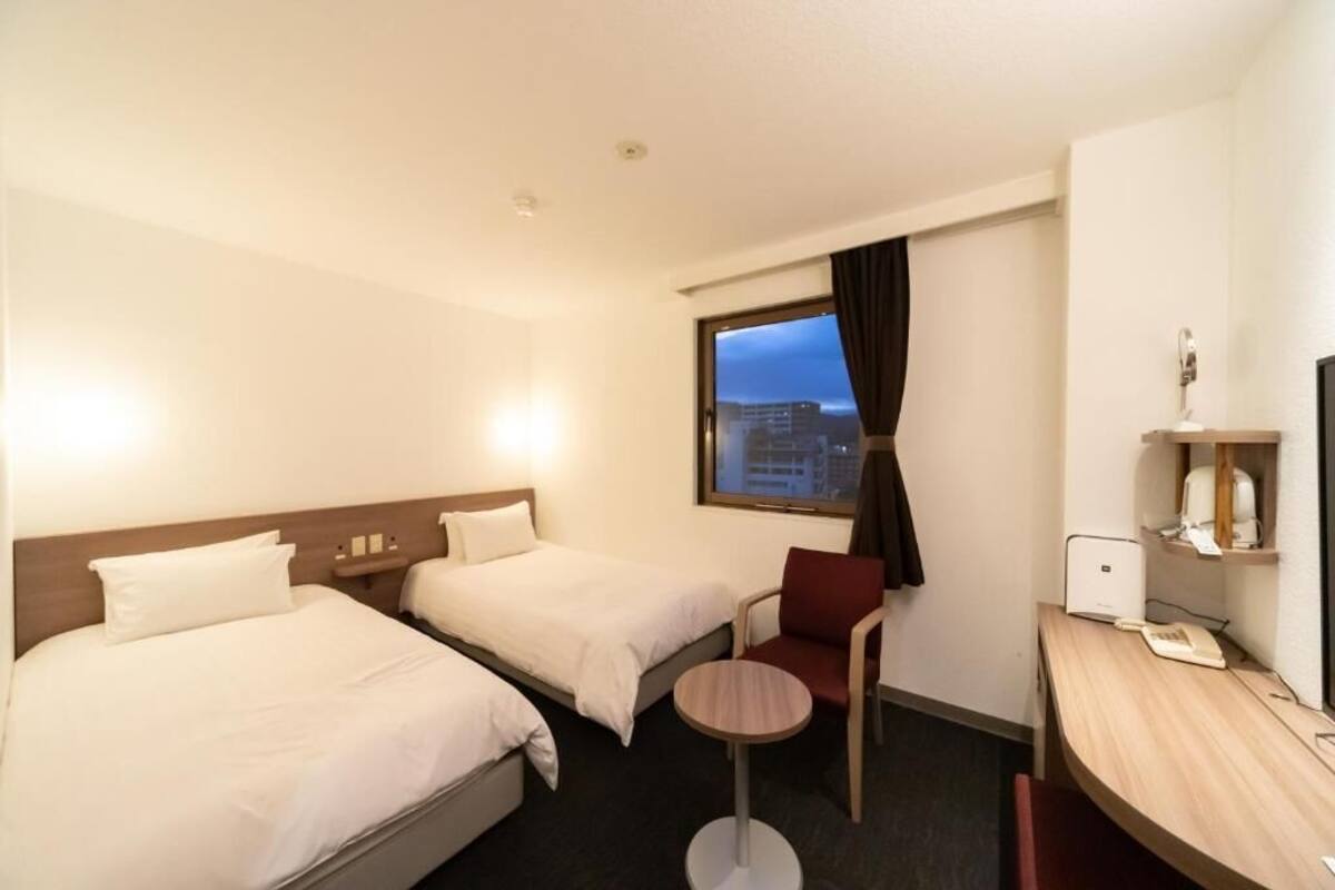 A hotel room with two beds, a desk, a chair, and a window with a view of buildings.