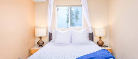 A neatly made bed with white bedding and a blue throw, two bedside lamps, and a window with a view of trees.