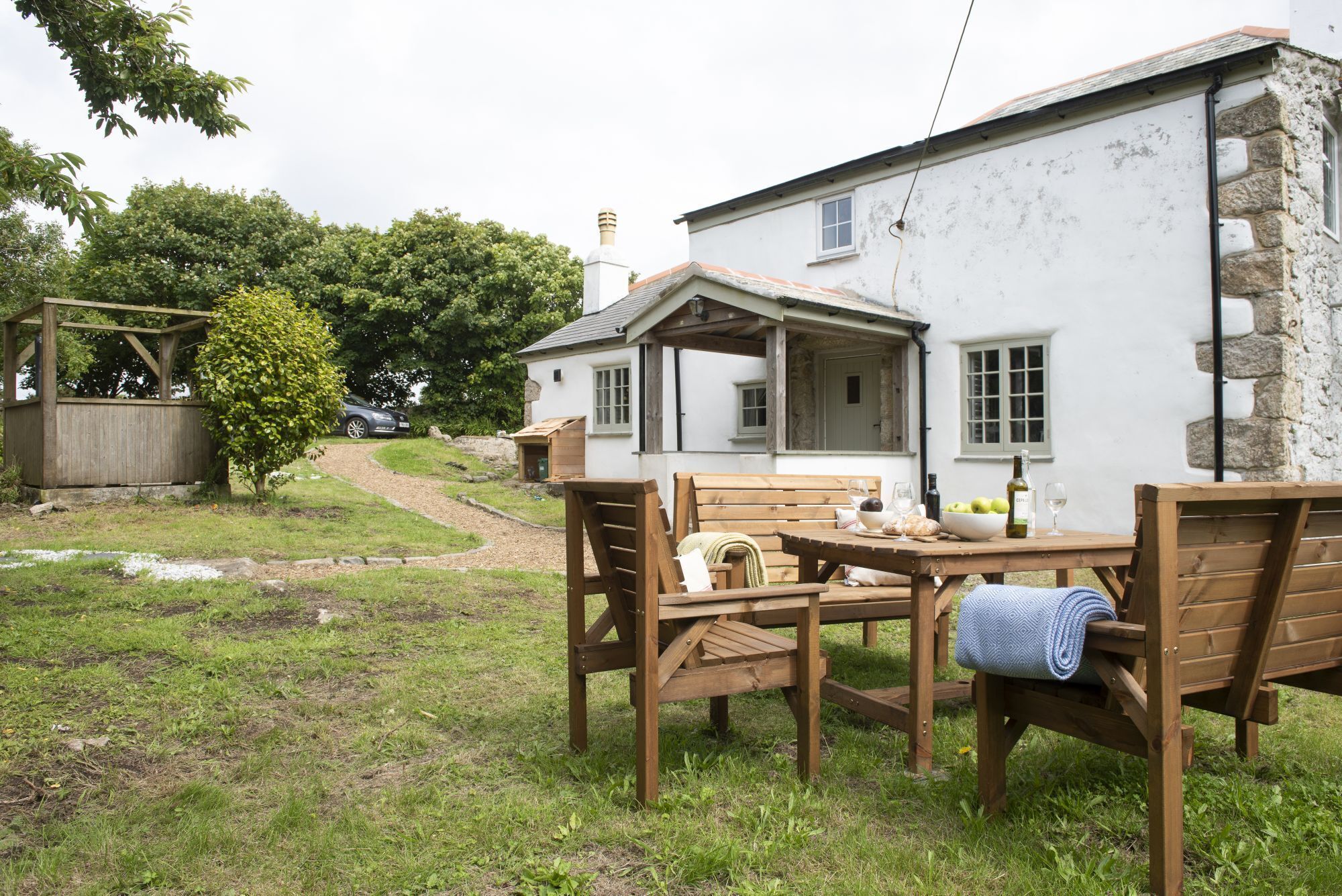 A rustic outdoor seating area with a wooden table and benches, a stone wall, and a white building in the background.
