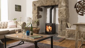 A cozy living room with a stone fireplace, a sofa, a coffee table with drinks and a book, and a rustic wooden side table. - Worvas Counthouse -  Country Cottage - Hot Tub - Pet Friendly (St Ives)