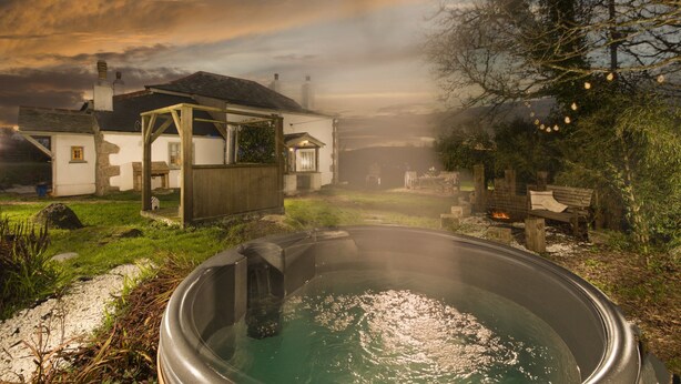 A rustic outdoor hot tub area with a wooden deck, a white building with a chimney, and a stone wall in the background. - Worvas Counthouse -  Country Cottage - Hot Tub - Pet Friendly (St Ives)