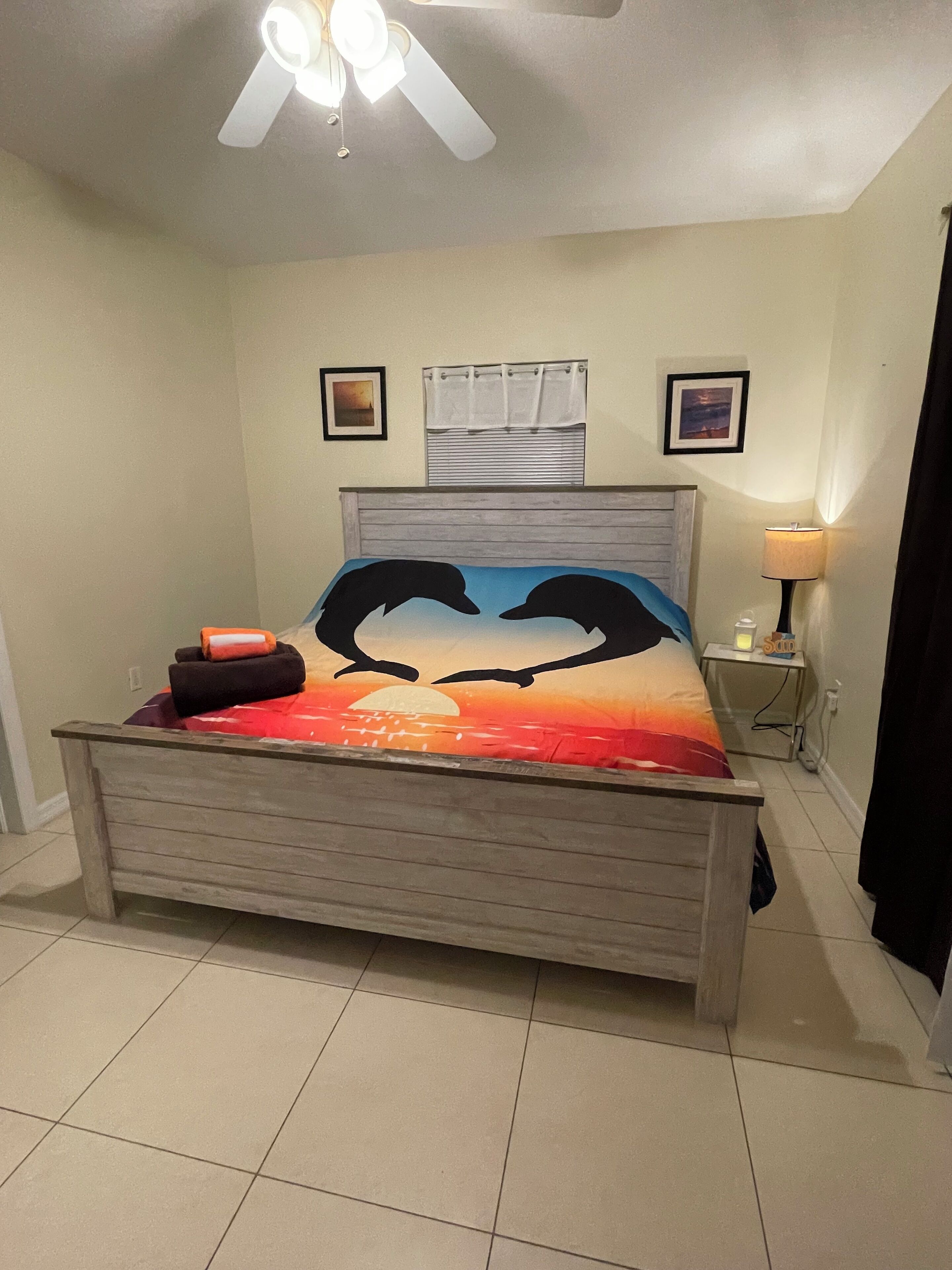 Ein Schlafzimmer mit einem Bett, das ein Design mit Sonnenuntergang und Delfinen zeigt, einem Deckenventilator, einem Nachttisch mit Lampe und zwei gerahmten Bildern an der Wand.