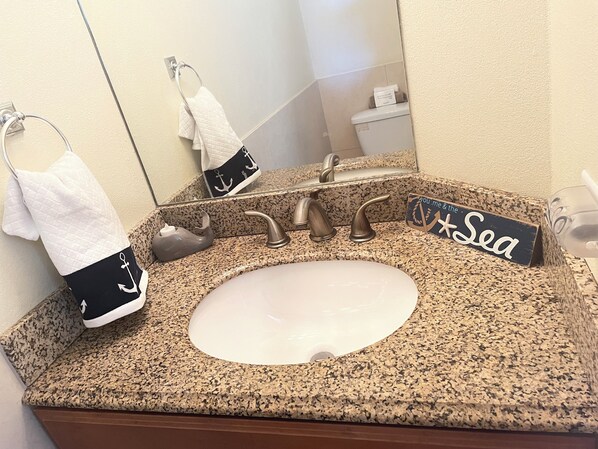 Ein Badezimmer mit einer Granit-Arbeitsplatte, einem Waschbecken und einem Spiegel. Handtücher hängen an einem Wäscheständer, und ein Schild trägt die Aufschrift 'you.me & the Sea'.