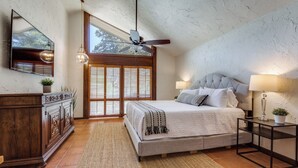 A bedroom with a large bed, a ceiling fan, a TV mounted on the wall, and a window with blinds.