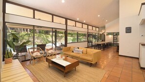A spacious living area with a sofa, chairs, and a coffee table. Large windows offer a view of a pool and greenery.