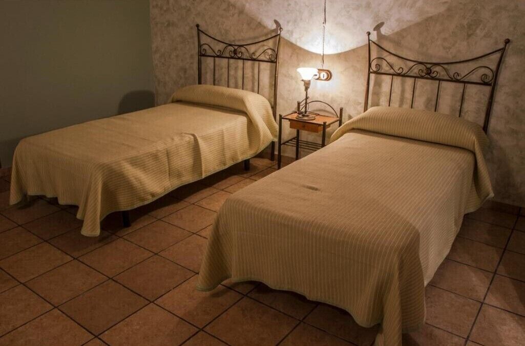 Dos camas individuales con cabeceras, una mesita de noche con lámpara, y suelo de baldosas.