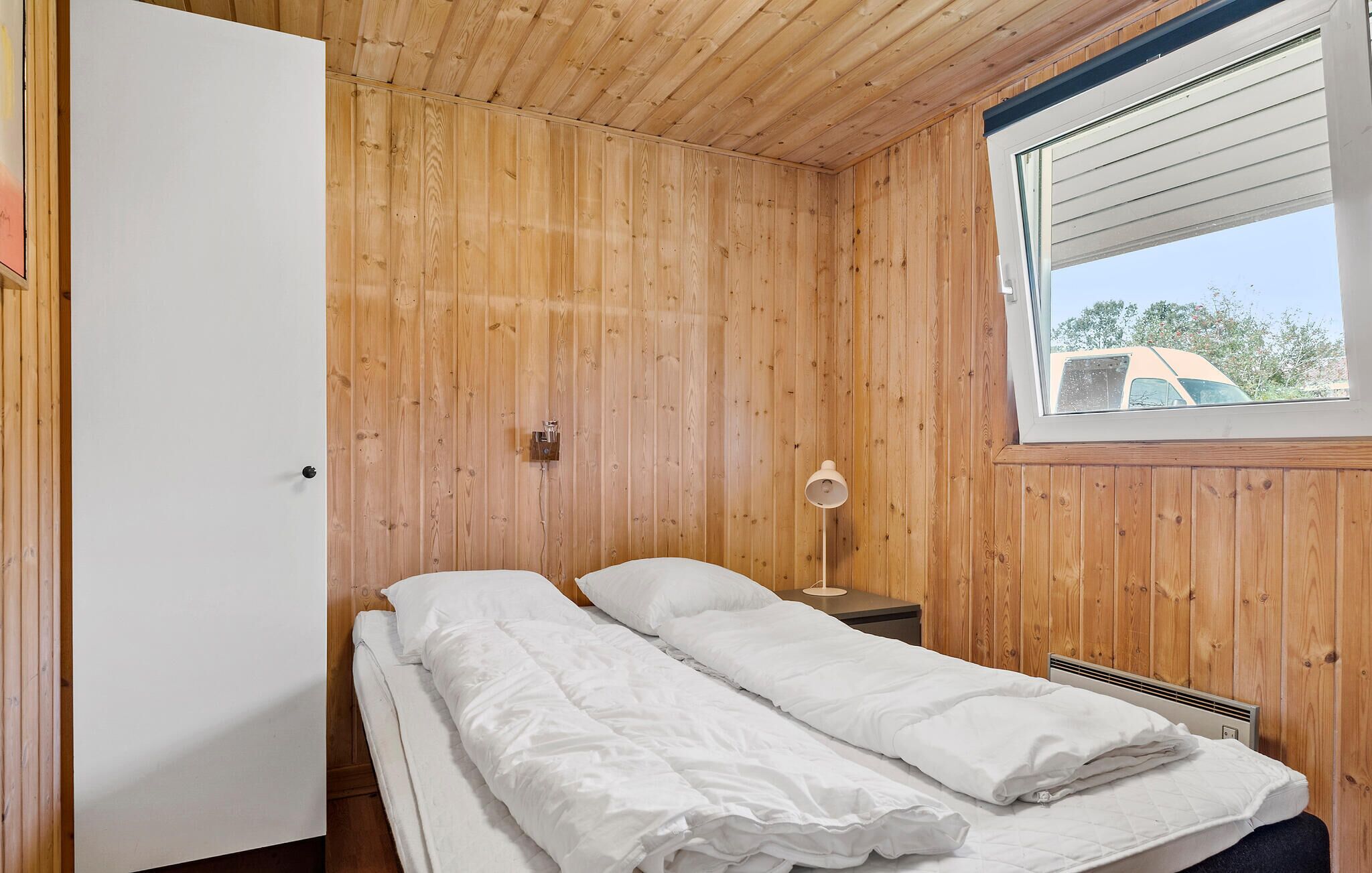 A small, wooden-paneled room with a bed, a nightstand, a lamp, and a window with a view of the outside.