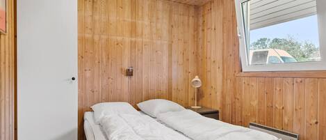 A small, wooden-paneled room with a bed, a nightstand, a lamp, and a window with a view of the outside.