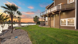 A two-story house with a balcony overlooking a lake, a sandy area with a palm tree, and a well-maintained lawn.