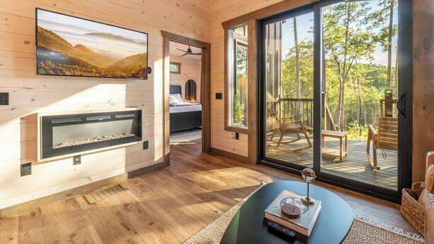 A cozy cabin interior with a fireplace, a large window, and a balcony with outdoor furniture. - New Modern Design! Treetop Escape! Hot Tub, Fireplace, Fire Pit & Views!! (Sevierville)