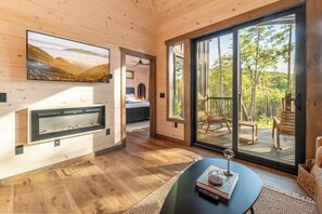 A cozy cabin interior with a fireplace, a large window, and a balcony with outdoor furniture. - New Modern Design! Treetop Escape! Hot Tub, Fireplace, Fire Pit & Views!! (Sevierville)
