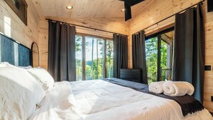 A bedroom with a large bed, dark curtains, and a view of trees through the windows. - New Modern Design! Treetop Escape! Hot Tub, Fireplace, Fire Pit & Views!! (Sevierville)