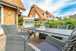 En udendørs terrasse med fletningsmøbler og et bord, omgivet af murstensbygninger og grøn vegetation.