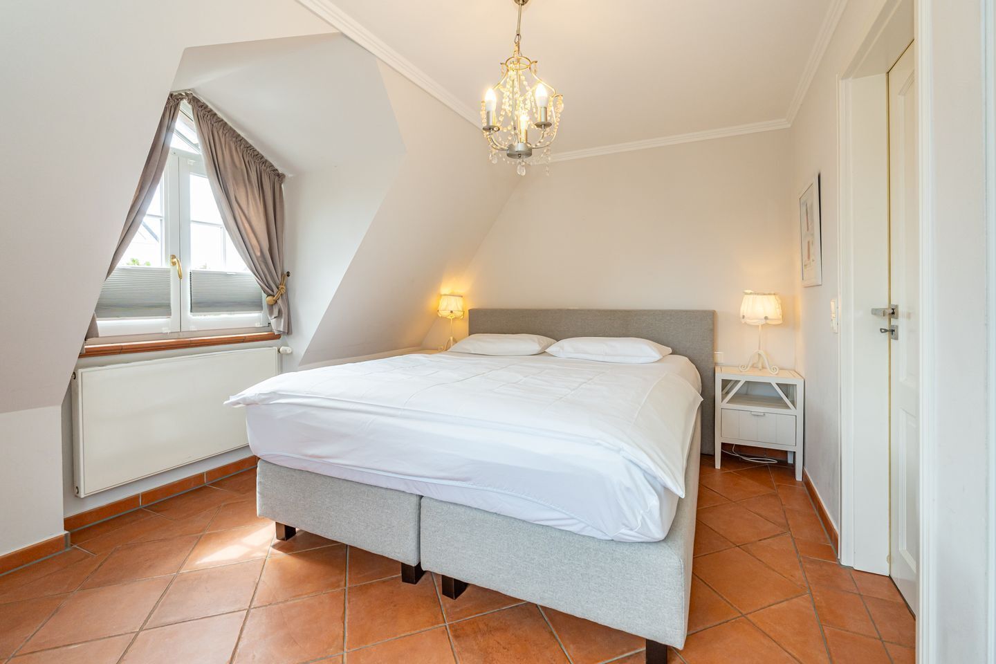 A bedroom with a bed, a chandelier, a nightstand, a window with curtains, and a radiator.